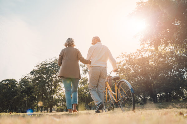 An older couple walking through a park as one pushes a bike.