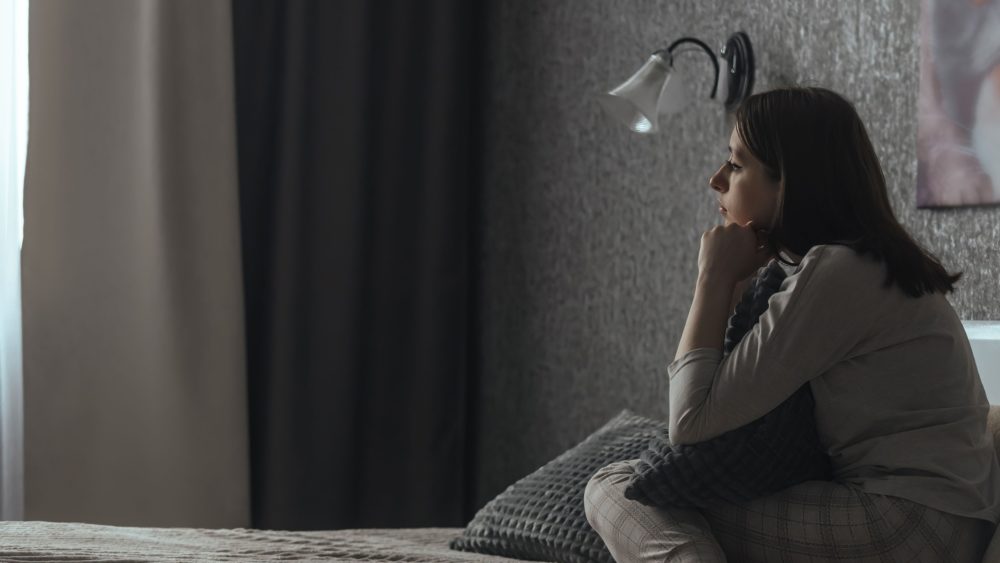 A woman sits in a dark bedroom looking sad.