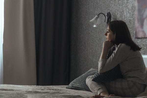 A woman sits in a dark bedroom looking sad.